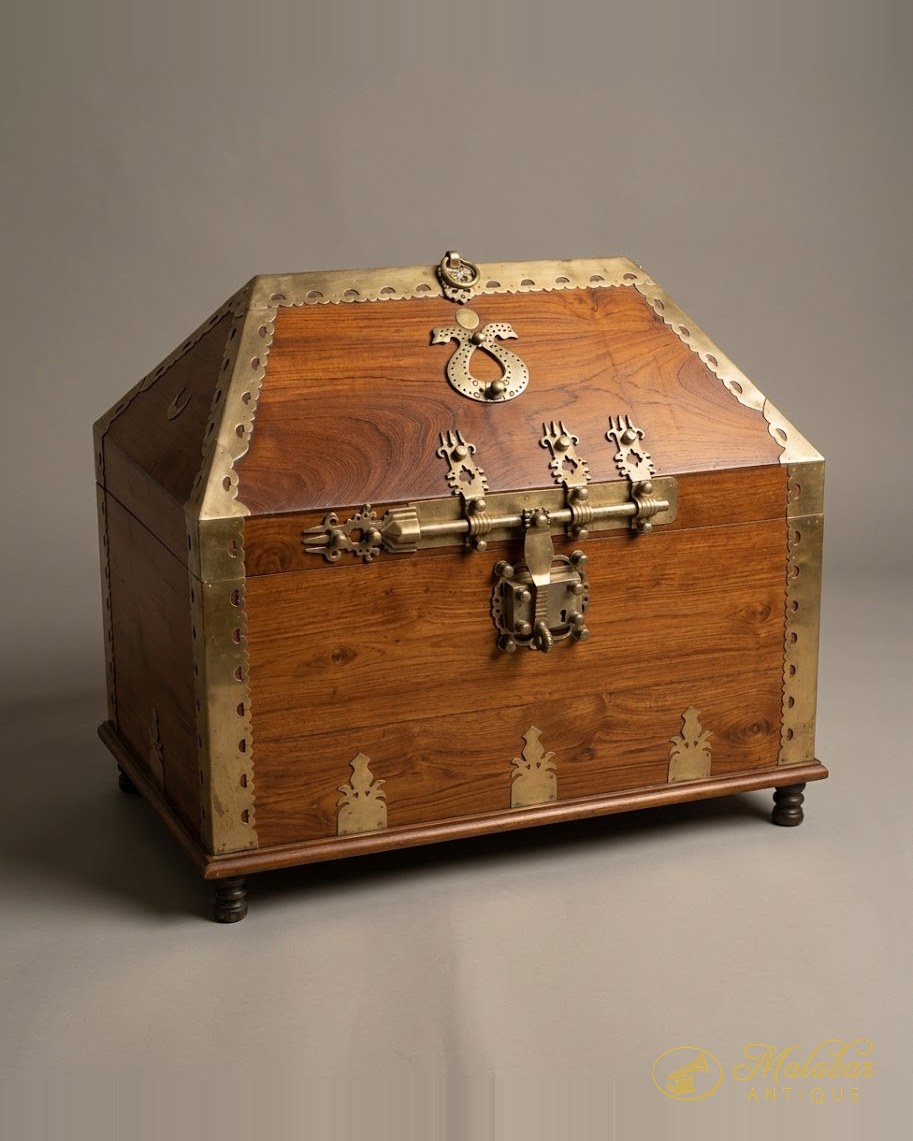 Large teak chest with a gabled lid, scalloped brass sheathing, a heavy brass latch bar, padlock plate and turned bun feet.