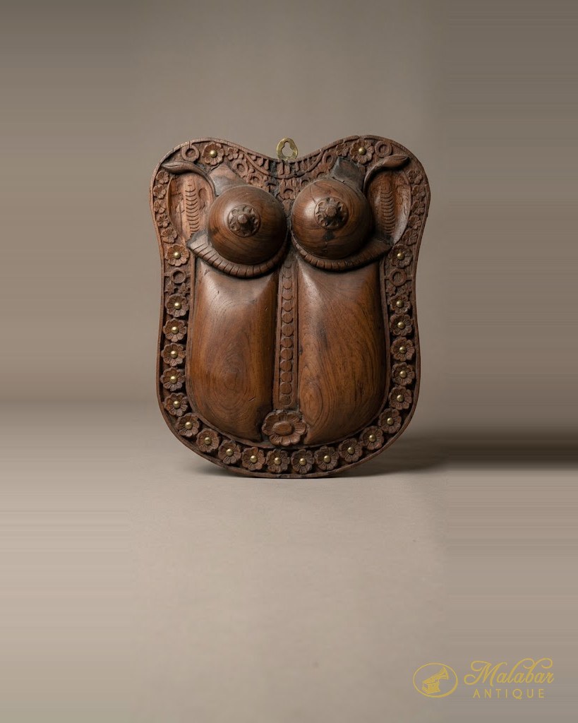 Shield-shaped teak wall plaque carved as a stylised female torso, the border ringed with carved rosettes and brass studs.