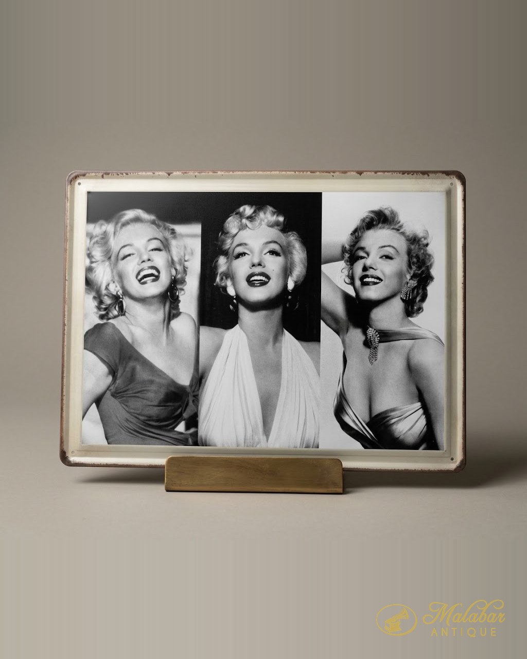 Enamelled tin sign with three black and white portraits of Marilyn Monroe, standing on a plain wooden display block.