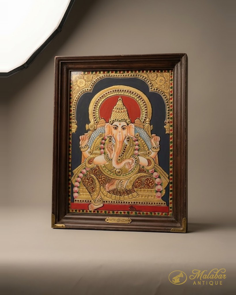Framed Tanjore painting of Ganesha seated beneath a gilded arch, worked in raised gold leaf with coloured stones on a deep blue ground.