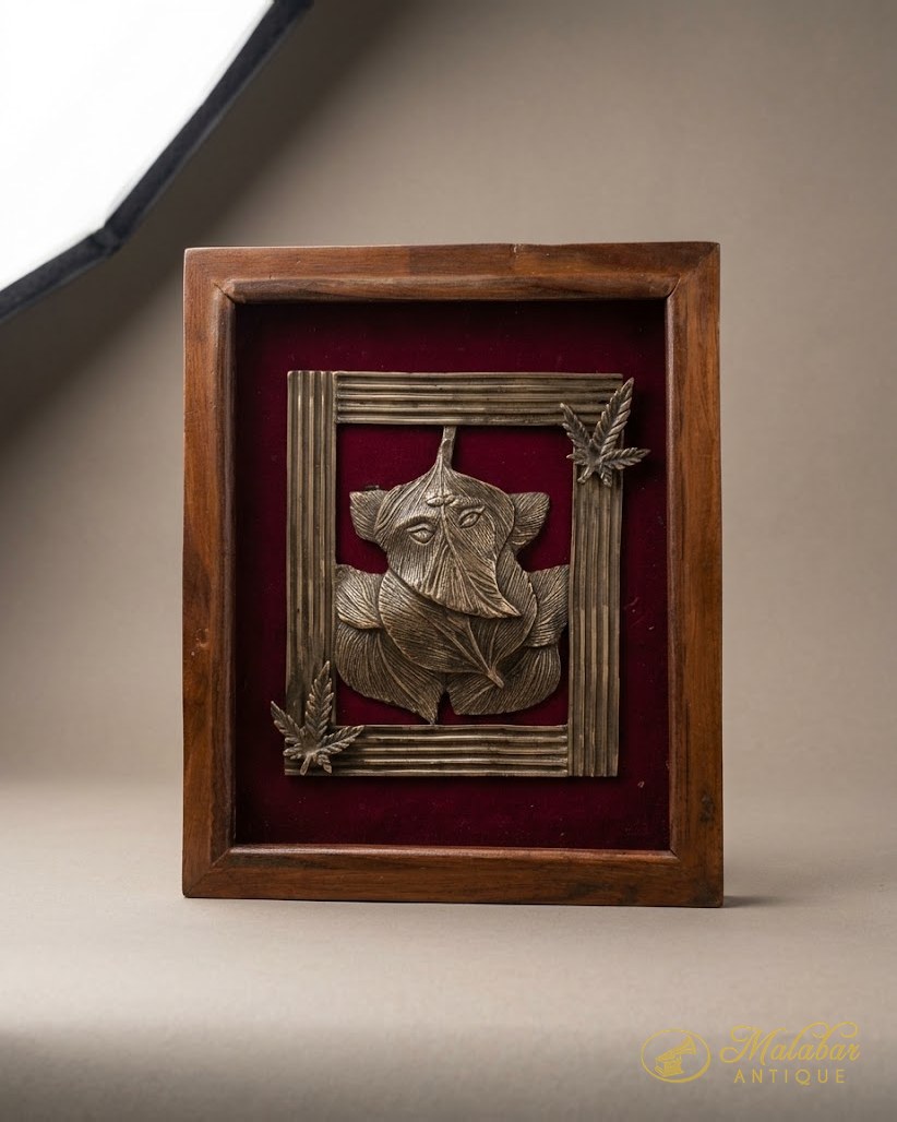 Cast brass Ganesha relief mounted on crimson velvet within a plain teak box frame, with brass leaf motifs at the corners.